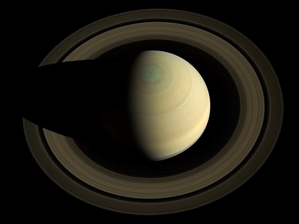 Saturn seen from above its north pole by the Cassini spacecraft — the pale yellow banded globe encircled by the broad concentric ring system, with the planet's shadow cutting a dark wedge across the rings
