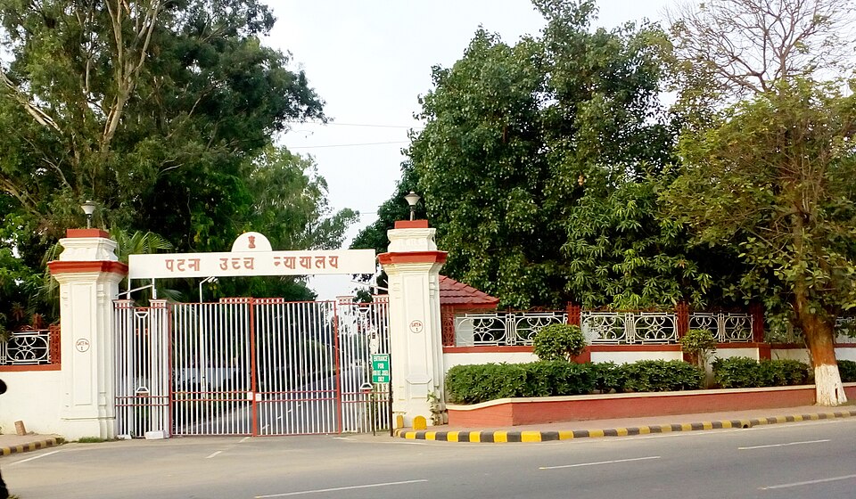 The street entrance to the Patna High Court: a white-and-red gateway with two pillars carrying the Hindi lettering 'पटना उच्च न्यायालय' above closed steel gates, flanked by a low ornamental boundary wall and large shade trees (the court building itself is not in frame)