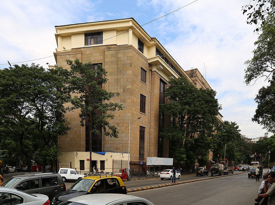 The Reserve Bank of India central office in Mumbai seen from the street corner: a severe stone-faced Art Deco block of five storeys with narrow recessed windows and a projecting cornice, street trees along its plinth and taxis and traffic on the road in front