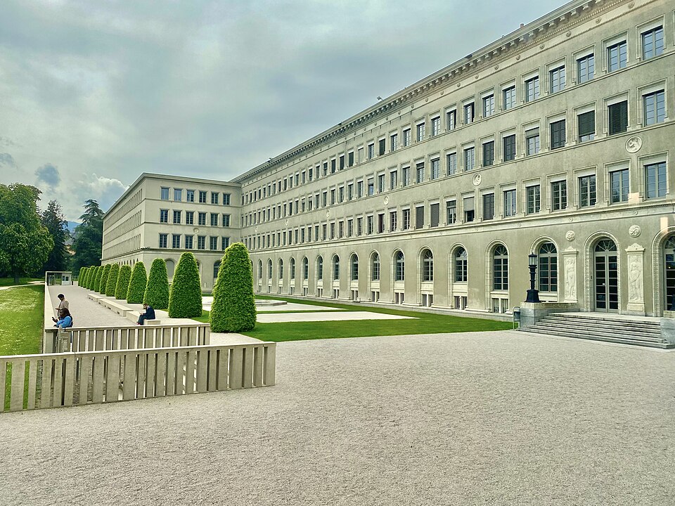 The Centre William Rappard in Geneva, home of the WTO: a long four-storey pale stone building with an arcaded ground floor and regular rows of windows, running away along a gravel forecourt with clipped conical shrubs and a lawn beside it