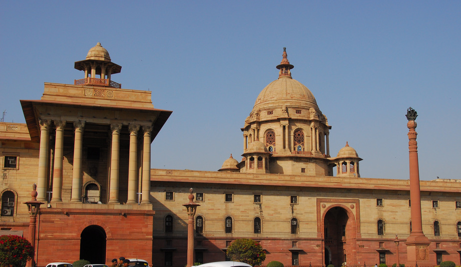 The North Block of the Secretariat on Raisina Hill: a long red and buff sandstone facade with a deep columned portico under a chhatri on the left, a large ribbed baroque dome on a windowed drum in the centre, an arched gateway below it and a tall carved column to the right