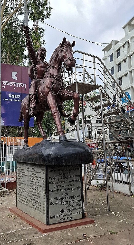 A dark bronze equestrian statue of Veer Kunwar Singh on a black plinth in a town square: a turbaned, bearded rider in a knee-length coat with his sword raised overhead, mounted on a horse with one foreleg lifted, the plinth carrying a long Hindi dedication