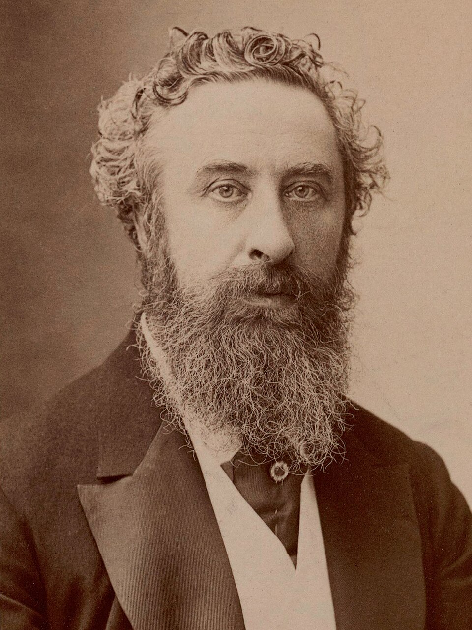 A sepia studio head-and-shoulders portrait of Lord Lytton by Nadar: a middle-aged man with tightly curled hair and a long straggling beard, in a dark jacket over a waistcoat with a stud and a pale sash across the chest