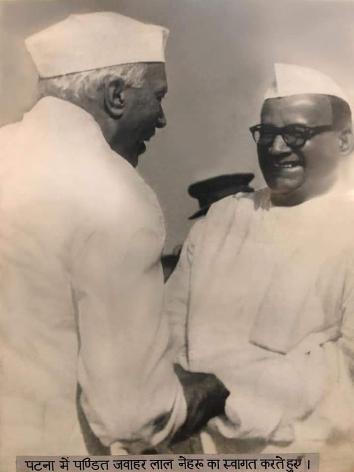 An archival black-and-white print showing Jawaharlal Nehru in profile, in a Gandhi cap, clasping hands with a smiling heavy-set man in thick spectacles, white kurta and Gandhi cap — Shri Krishna Sinha — with a printed Hindi caption strip below reading 'पटना में पण्डित जवाहर लाल नेहरू का स्वागत करते हुए।'