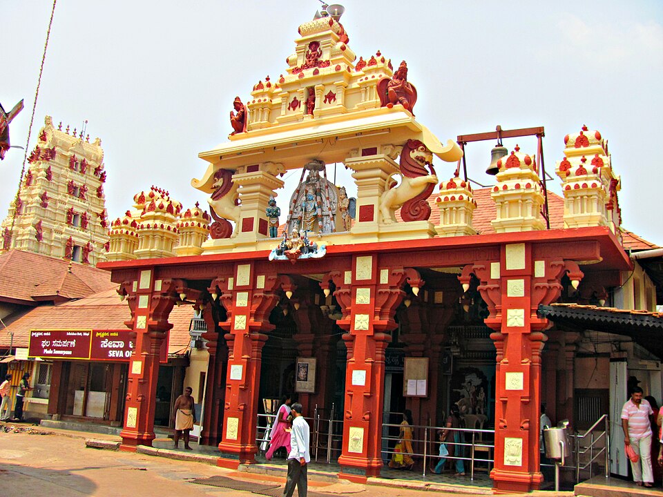 उडुपी के श्री कृष्ण मठ का प्रवेश-द्वार: लाल और क्रीम रंग का स्तंभयुक्त बरामदा, जिसके ऊपर गोपुरम शैली की मंज़िलदार संरचना है जिस पर देवताओं तथा एक उछलते सिंह की पलस्तर-मूर्तियाँ हैं, साथ में लटकती पीतल की घंटी, कन्नड़ में लिखे सूचना-पट और अगले आँगन में दर्शनार्थी