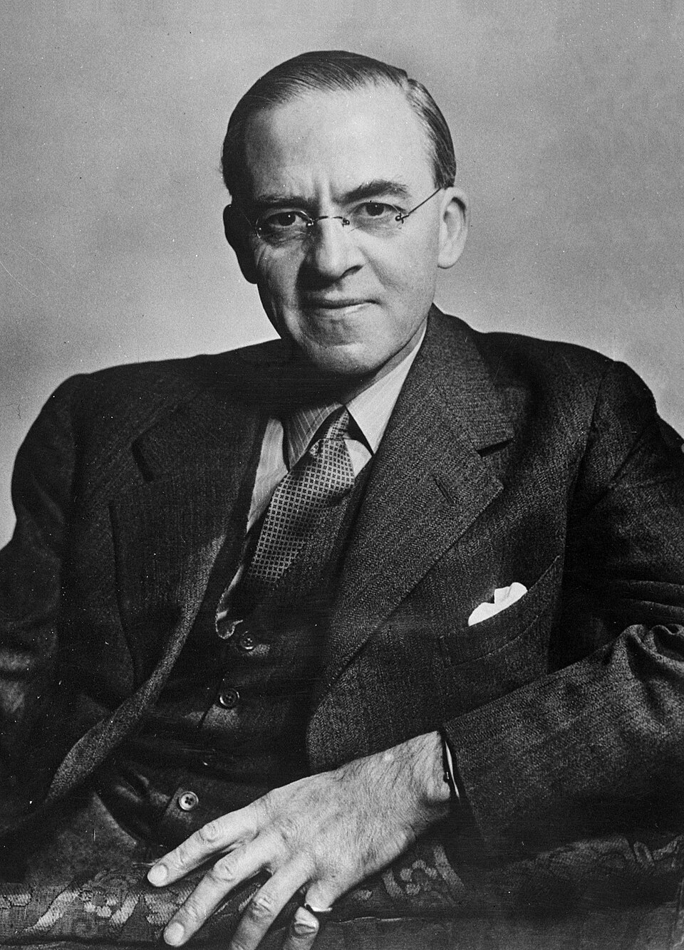 A 1947 black-and-white studio portrait of Sir Stafford Cripps: a balding, thin-faced man in rimless spectacles, seated in a tweed suit with waistcoat and patterned tie, one hand resting on the arm of an upholstered chair