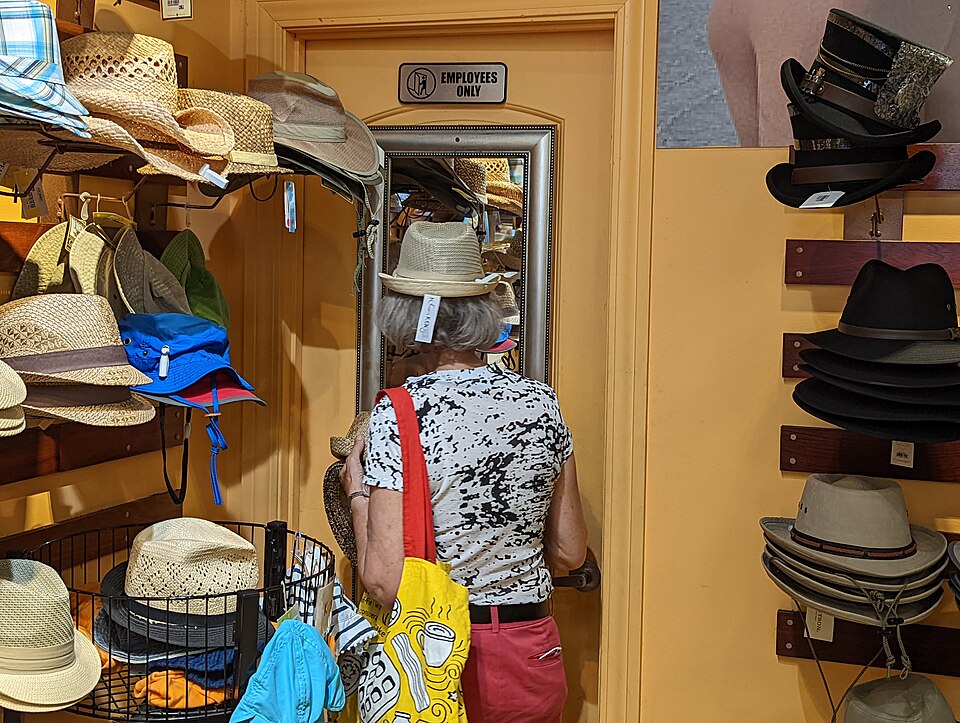 Inside a hat shop: straw trilbies, sun hats and felt fedoras stacked on wall brackets and wire racks on both sides of a narrow yellow-walled aisle, with a customer seen from behind trying a pale straw hat in a wall mirror