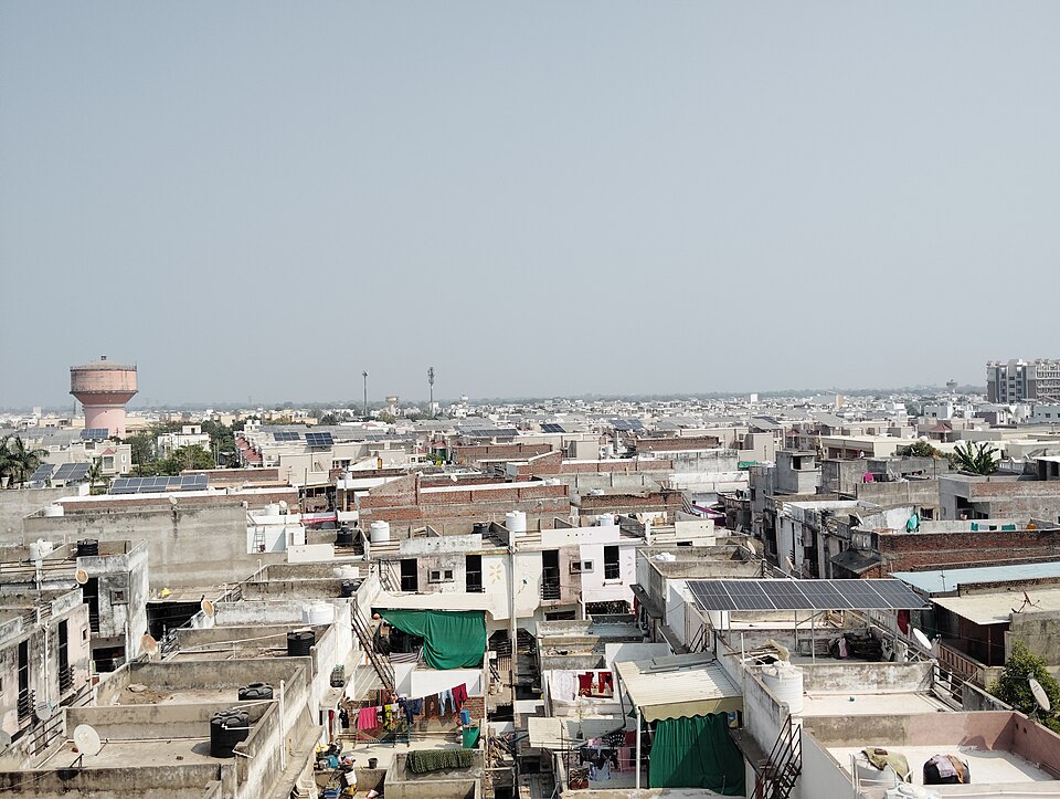 A view across the flat concrete rooftops of a dense Indian town (Mehsana, Gujarat), with blue photovoltaic panel arrays mounted on several of the roofs among the black water tanks, dish antennas and drying laundry