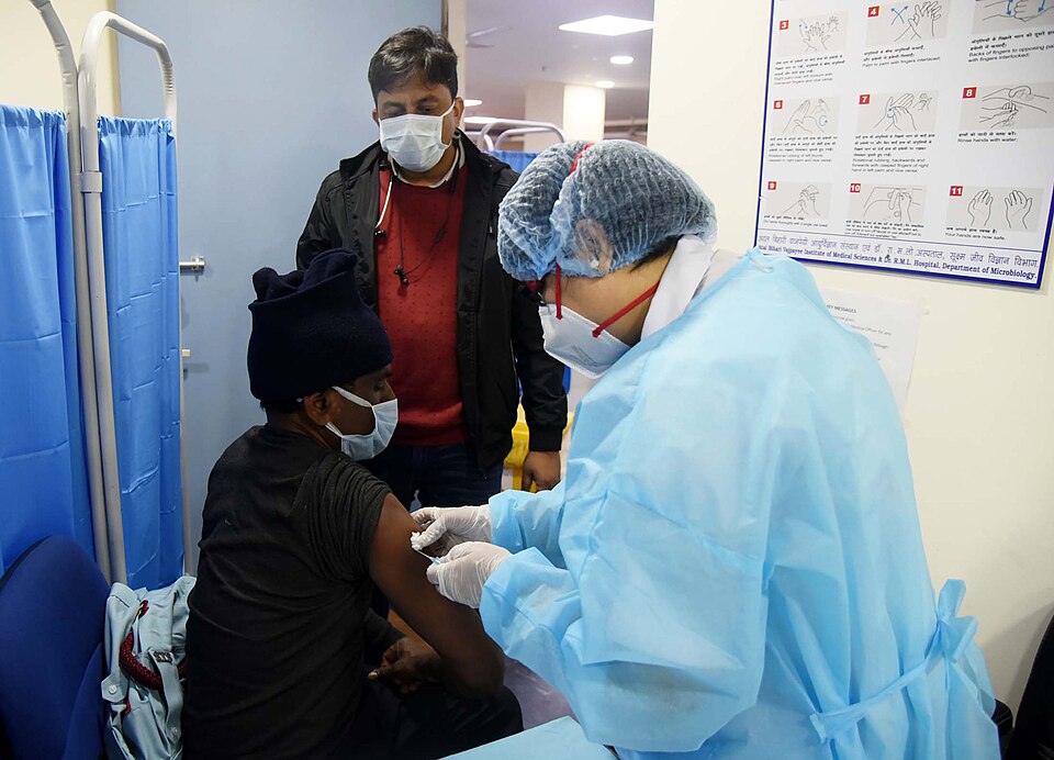 A health worker in a blue PPE gown, hair cap, N95 mask and gloves injecting a COVID-19 vaccine into the upper arm of a seated masked man at a Delhi hospital, with a masked doctor looking on and a Hindi-English handwashing poster on the wall
