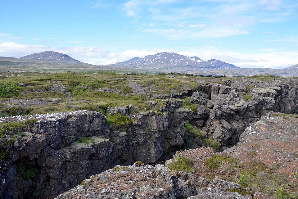 A deep open fissure splitting the layered basalt bedrock in the foreground at Þingvellir, Iceland, with the moss-and-grass rift plain stretching away to snow-streaked hills under a blue sky