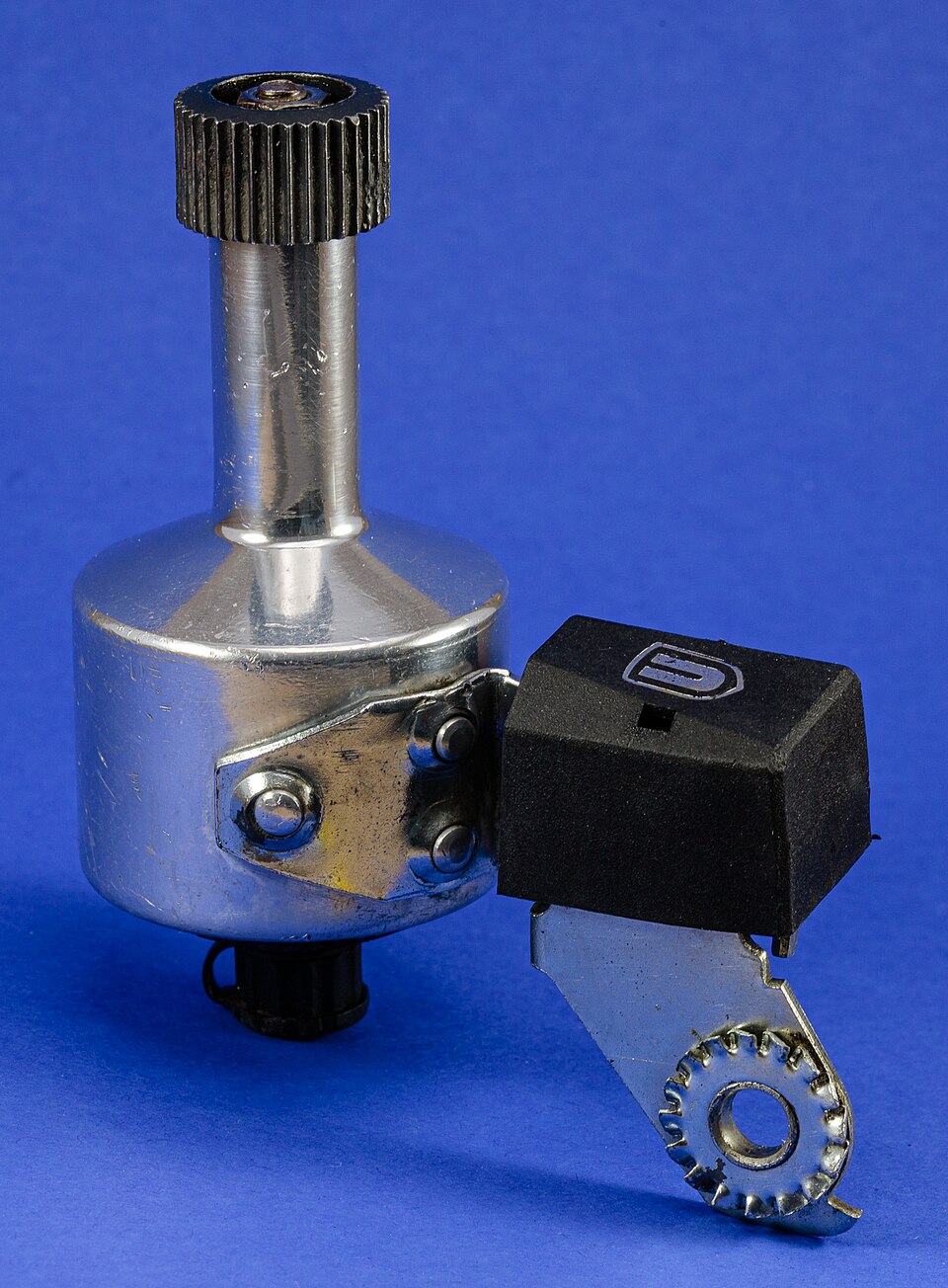 A chrome bicycle bottle dynamo photographed on a blue background — the cylindrical generator body with its knurled black drive roller on top, alongside the hinged steel mounting bracket and toothed clamp that fix it to the fork