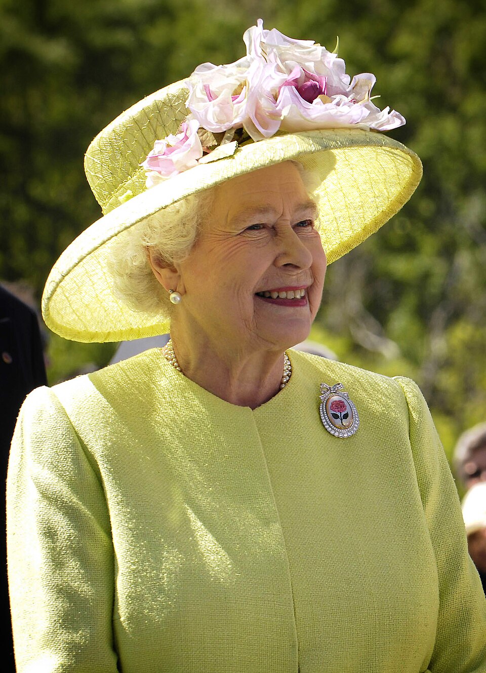 Queen Elizabeth II photographed outdoors in 2007, smiling in profile, wearing a lime-yellow coat and matching wide-brimmed hat trimmed with pink and white flowers, pearl necklace and earrings and a rose brooch