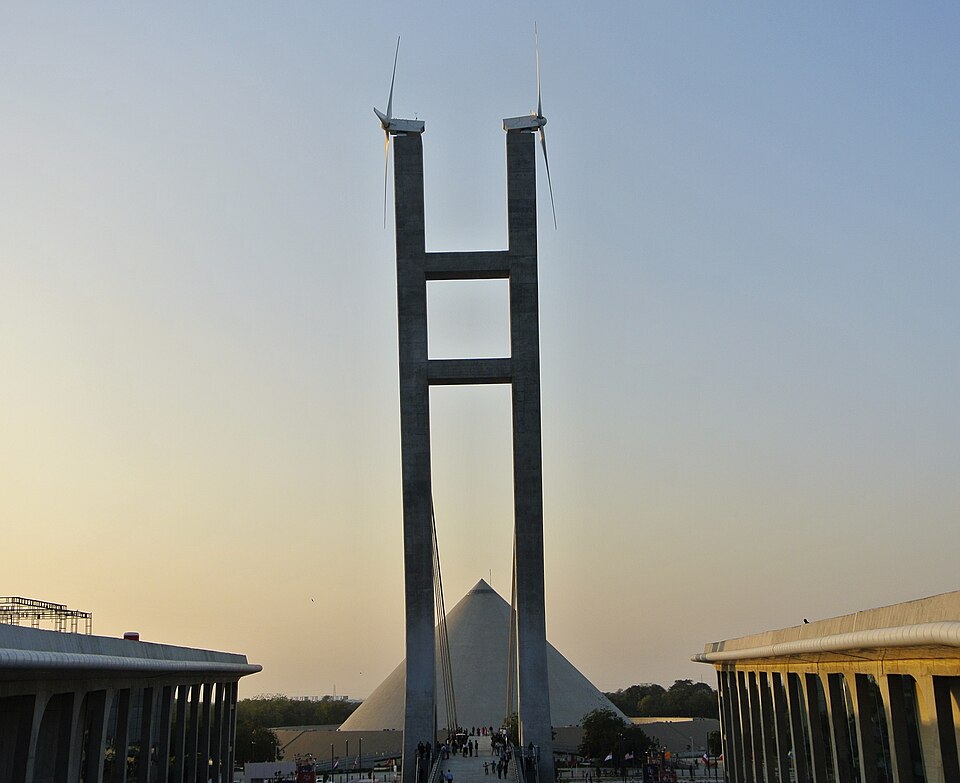 The Mahatma Mandir complex at Gandhinagar at dusk — the twin concrete pylons of its landmark gateway carrying wind-turbine blades, the pyramidal Dandi Kutir museum rising behind, and the long colonnaded convention halls flanking the plaza on both sides