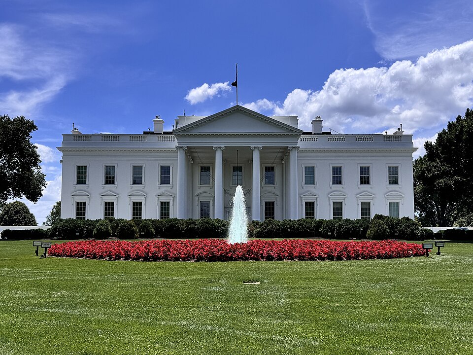 The north front of the White House in Washington DC — the white neoclassical facade with its columned North Portico and pediment, the flag on the roof, and the North Lawn fountain ringed by a bed of red flowers in the foreground