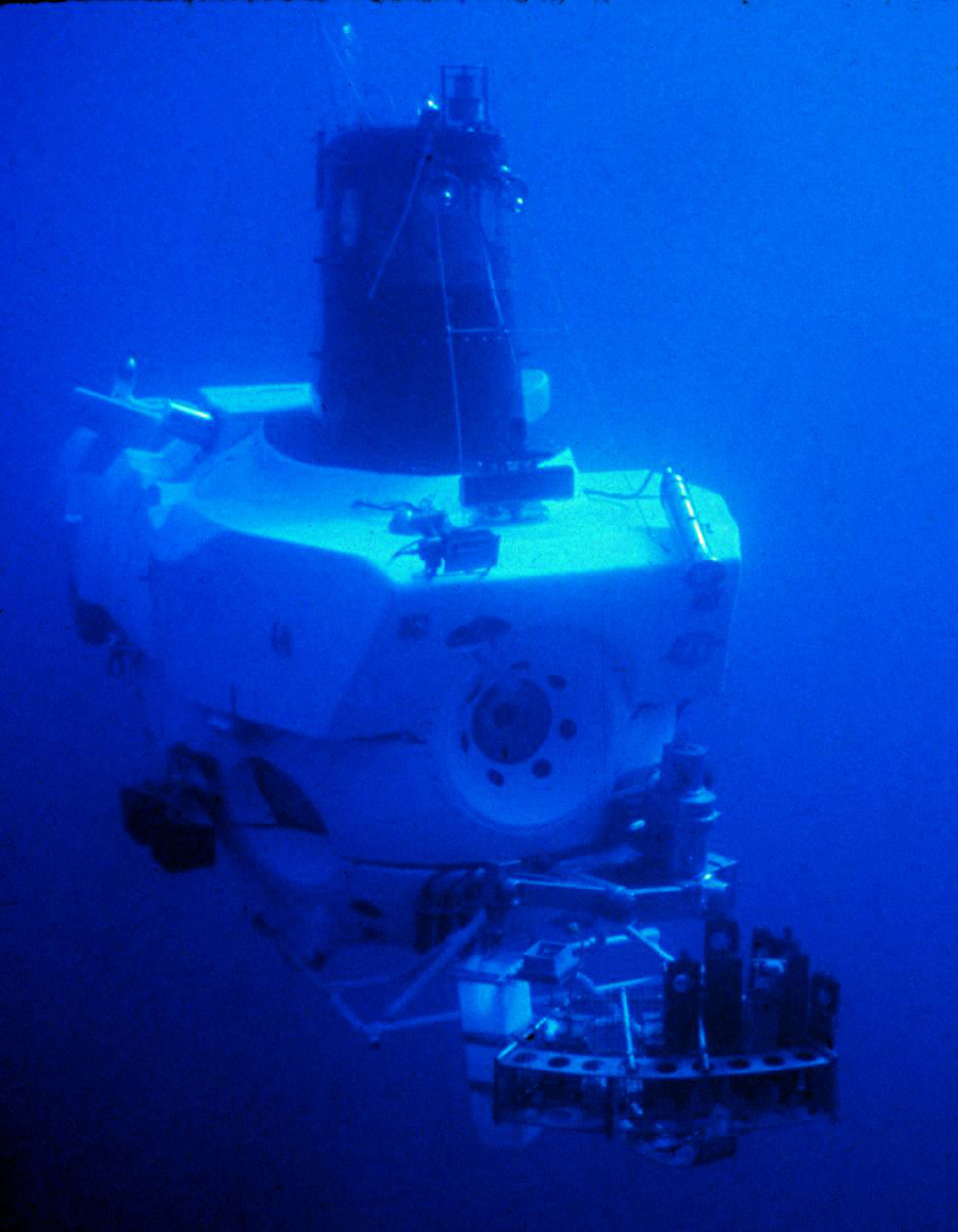 The white deep-sea submersible Alvin photographed underwater in deep blue water — its blocky hull and conning tower above, with the manipulator arm and the instrument-and-sample basket slung at the bow