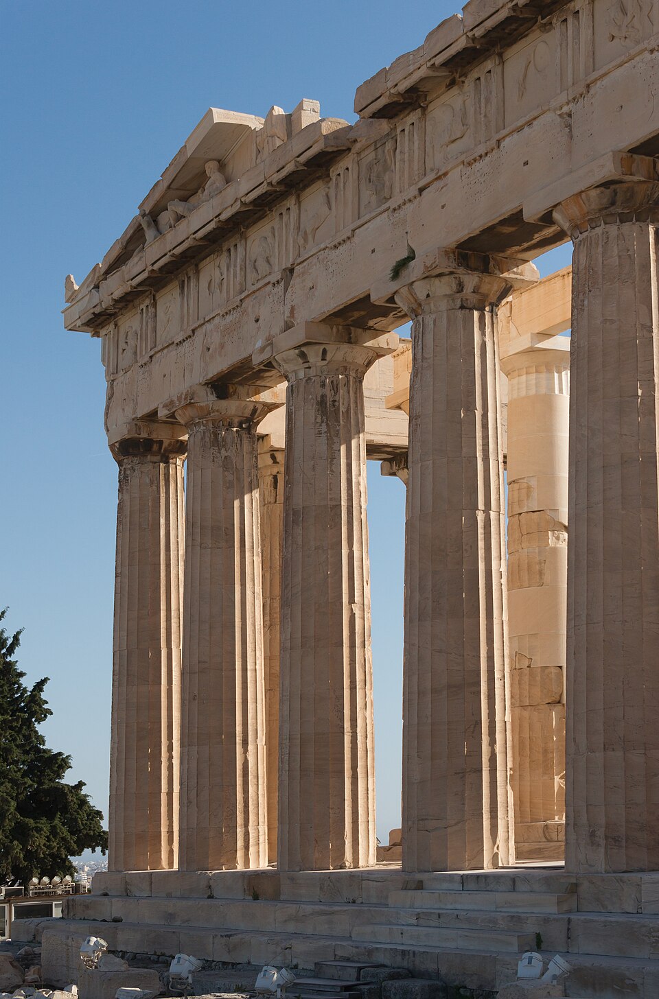 The east front of the Parthenon in low sunlight — a row of fluted Doric columns carrying the architrave, triglyphs and the sculpted pediment, with the newer pale restored drums of one column visible behind