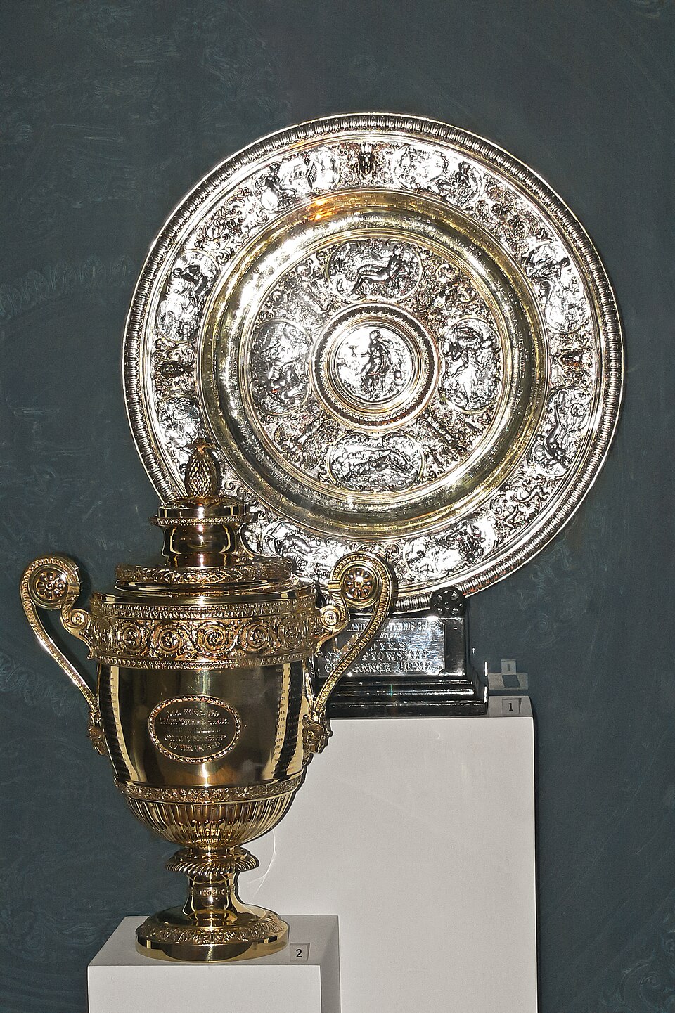 The Wimbledon trophies on display against a dark wall — the large circular Venus Rosewater Dish, its silver-gilt face worked in repoussé with mythological figures in a central roundel and surrounding cartouches, standing behind the gold men's singles cup on its plinth