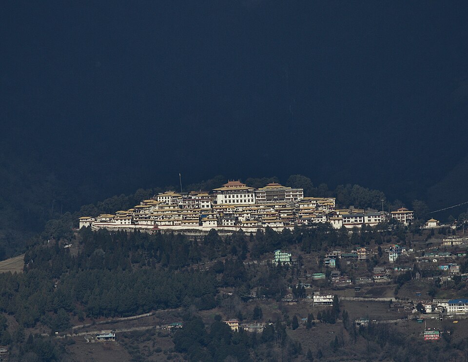 Tawang Monastery seen across the valley — a tightly packed complex of white buildings with gold-and-red roofs spread along a ridge top, dark forested mountainside rising behind it and the scattered houses of Tawang town on the slope below