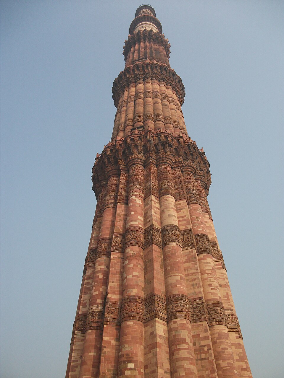 The Qutb Minar photographed looking up from its base — the tapering red-and-buff sandstone shaft with alternating angular and rounded flutes, ringed by carved calligraphic bands and three corbelled balconies, against a clear sky