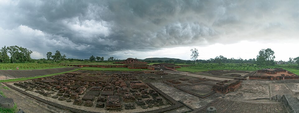 Panorama of the excavated Vikramshila site at Antichak under a storm sky — dozens of square brick votive-stupa bases laid out in rows in the foreground and the terraced brick platform of the main stupa on its raised plinth behind, with the monastery walls running off to the right