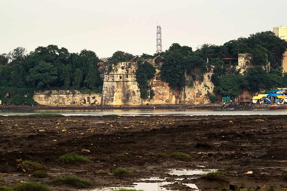 Surat Castle seen across the Tapi at low tide — the old fort's whitewashed rounded bastion and stretch of riverside curtain wall, heavily overgrown with trees, with the exposed mudflats of the river bed in the foreground