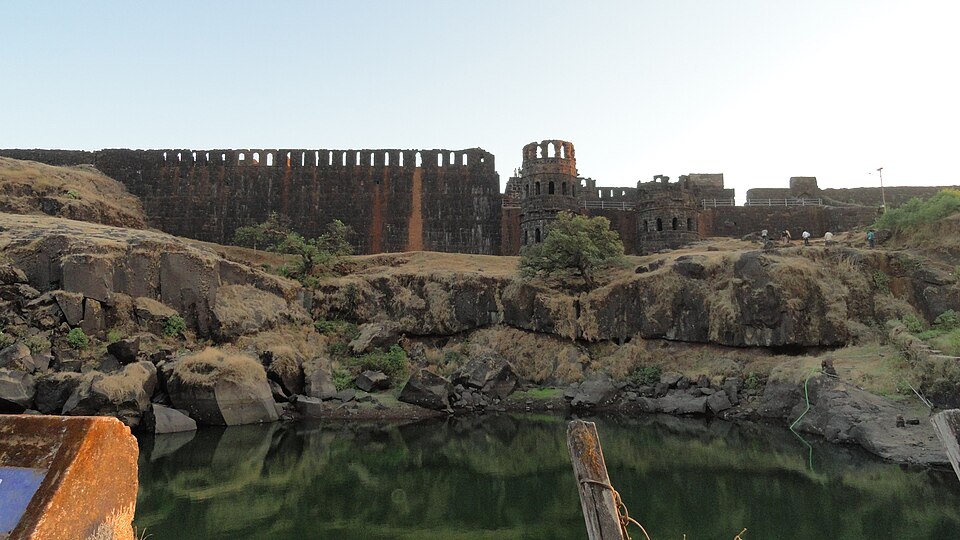 Raigad Fort in Maharashtra — a long dark stone rampart pierced by a row of arched openings running along the hill top, with a ruined multi-storeyed gate tower beside it, the bare rock of the plateau below and a green water cistern in the foreground