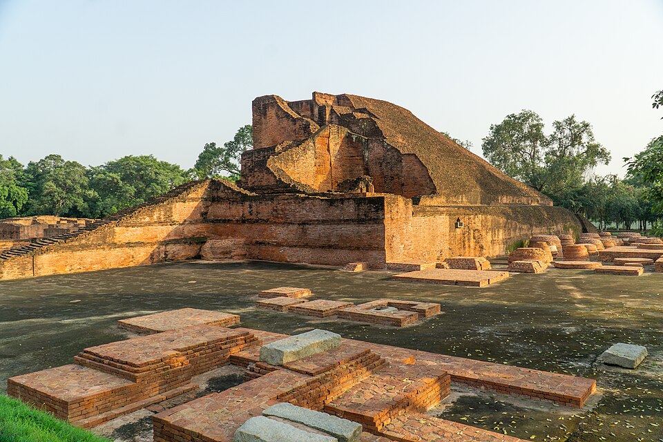 The excavated Nalanda Mahavihara at sunrise — the great brick mass of the Sariputta stupa (Temple 3) rising in stepped terraces on its plinth, ringed by rows of small brick votive stupas and the low plinths of the surrounding monastery buildings