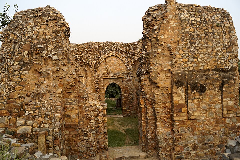 The roofless rubble-masonry chamber of Balban's tomb in Mehrauli Archaeological Park — thick walls of undressed stone with a pointed true arch framing the doorway and a second arched recess beside it, open sky above and grass showing through the opening