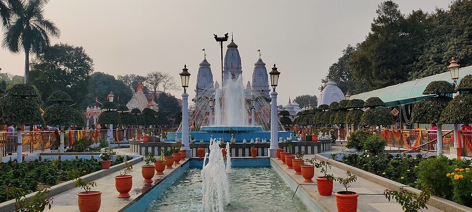 The garden forecourt of the Gorakhnath Temple at Gorakhpur: a tiered fountain playing in a blue-tiled pool, with the temple's three white shikharas and flagstaffs rising behind the trees and lamp posts