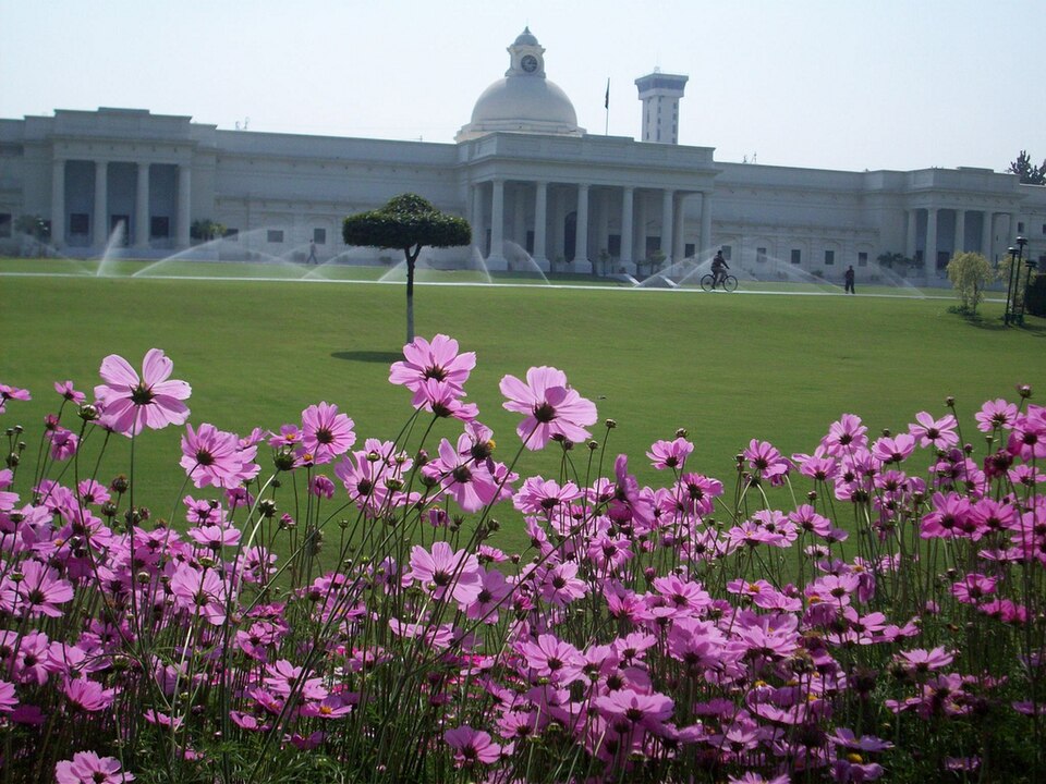 The long white colonial main building of IIT Roorkee seen across its lawn — a central dome and clock over a columned portico, flanked by colonnaded wings, with sprinklers running on the grass and a bank of pink cosmos flowers in the foreground