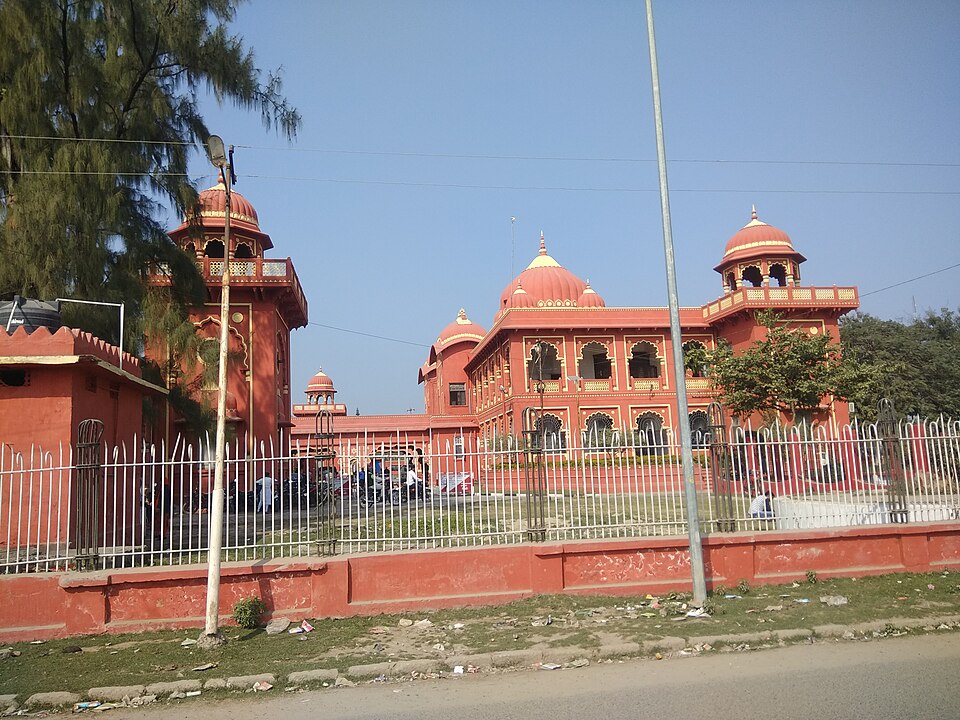 The Darbhanga Raj Kila seen from the road: a two-storey terracotta-red palace block with arched verandahs, ribbed domes and domed corner pavilions, standing behind a low boundary wall and iron railing