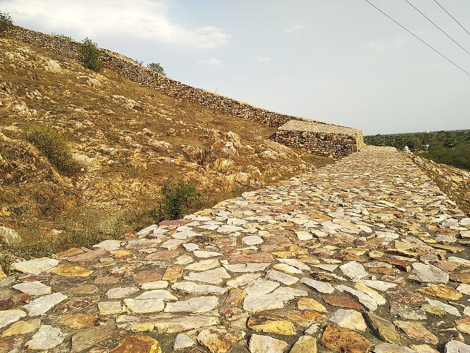 The Cyclopean Wall at Rajgir: a broad rampart of undressed stone blocks laid without mortar, its paved top running away along a bare hillside and continuing as a low stone crest over the ridge behind
