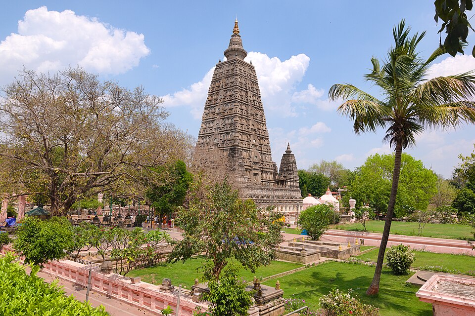 The Mahabodhi Temple at Bodh Gaya: the tall straight-sided brick tower, tiered with niched panels and capped by a gilded finial, rising above a smaller corner shrine, garden lawns, palms and a low pink boundary wall