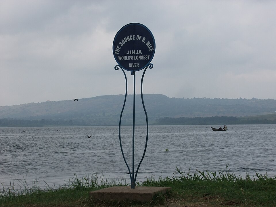 A round blue marker plaque on the grassy shore at Jinja reading 'THE SOURCE OF R. NILE - JINJA - WORLD'S LONGEST RIVER', with the open grey water of Lake Victoria, a fisherman's boat, wheeling birds and a low far shore behind it