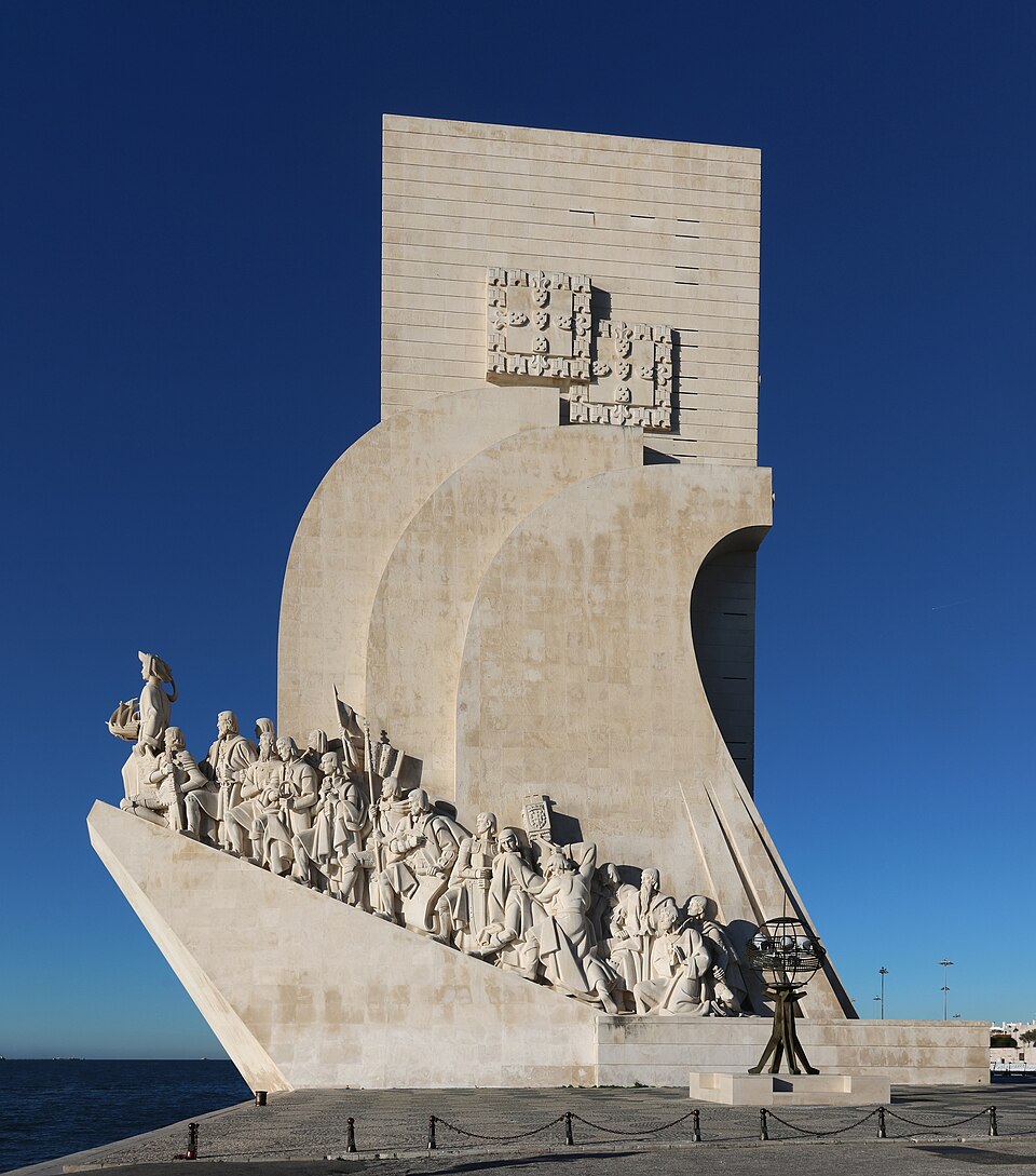 The Padrao dos Descobrimentos at Belem, Lisbon: a pale limestone monument shaped like a caravel's prow rising from the Tagus quayside, its sloping edge carved with a file of standing navigators, explorers and monks led by a figure holding a ship