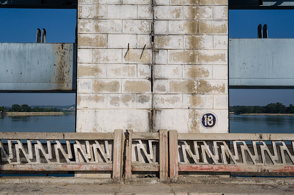 A close view along the deck of the Gandak Barrage: a whitewashed concrete pier marked '18' with a vertical joint down its centre, raised steel sluice gates on either side and the wide Gandak river with its far wooded bank showing through the openings