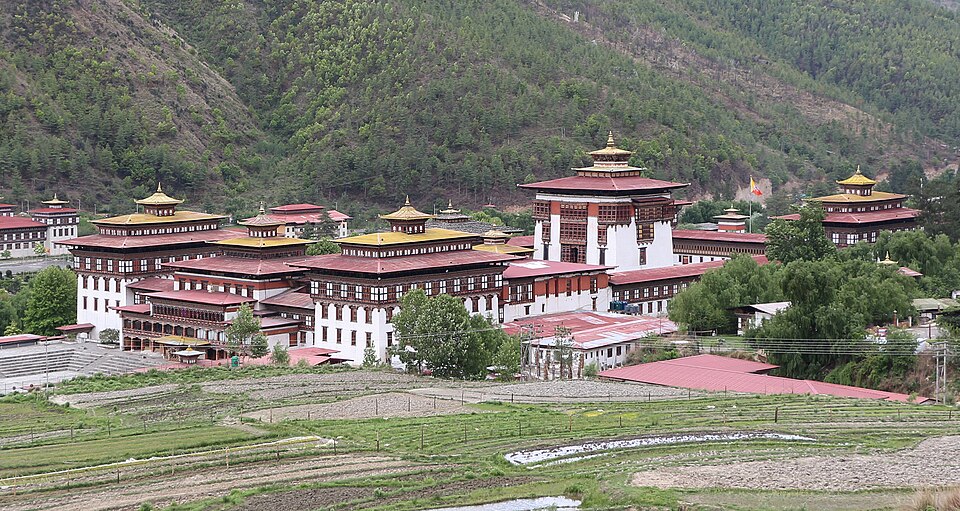 Tashichho Dzong at Thimphu seen from across the valley: a large whitewashed Bhutanese fortress-monastery with red-banded walls, tiered red roofs and gilded pinnacled towers, backed by a steep conifer-covered hillside and fronted by terraced fields
