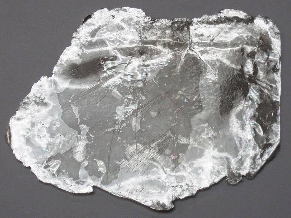 A single split sheet of muscovite mica lying on a grey background: a thin, silvery, near-transparent flake with an irregular outline, its surface showing the layered peeling and faint iridescent sheen of the cleaved basal planes
