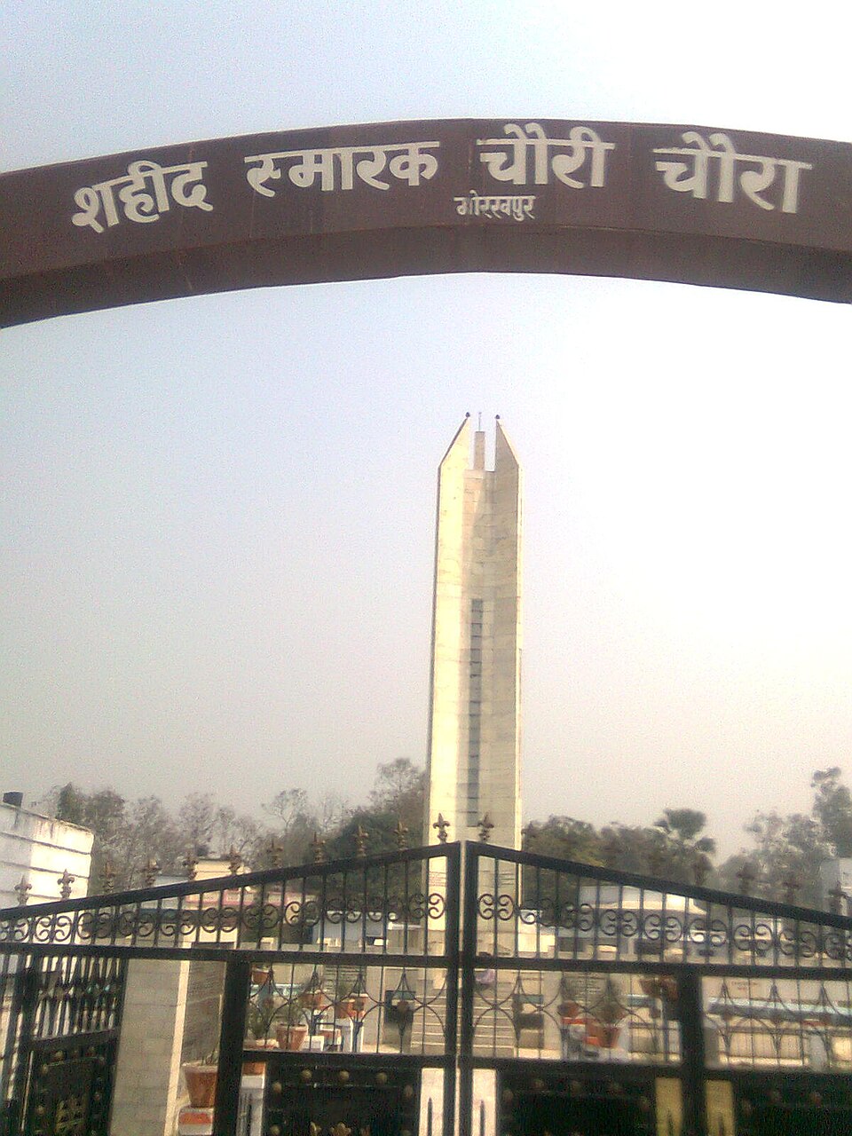 Looking through the entrance of the Chauri Chaura memorial: a brown steel arch overhead lettered in Hindi 'शहीद स्मारक चौरी चौरा, गोरखपुर', black wrought-iron gates below, and beyond them a tall pale stone obelisk with a stepped, twin-pointed top rising above bare winter trees in a white haze.
