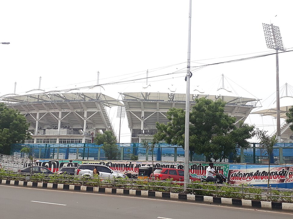 The Chepauk stadium seen from the road outside: three white tiered stands under sweeping cantilevered tensile roofs held up by raking steel struts, a lattice floodlight tower at the right, blue site hoarding and Tamil-lettered advertising boards along the boundary wall, with parked cars, a motorcyclist and a planted road divider in the foreground.