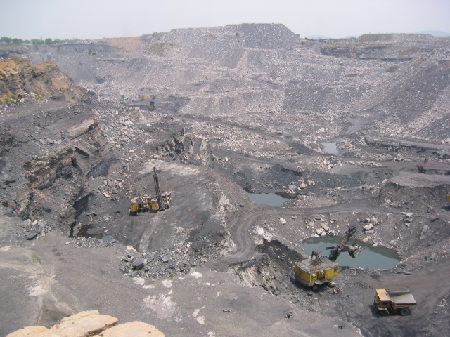 A wide opencast pit in the Jharia coalfield: grey and black rock benches stepping down to the floor of the working, hauling ramps cut into the sides, three pools of standing green water, and a yellow drilling rig, a shovel and a dump truck small against the scale of the excavation.