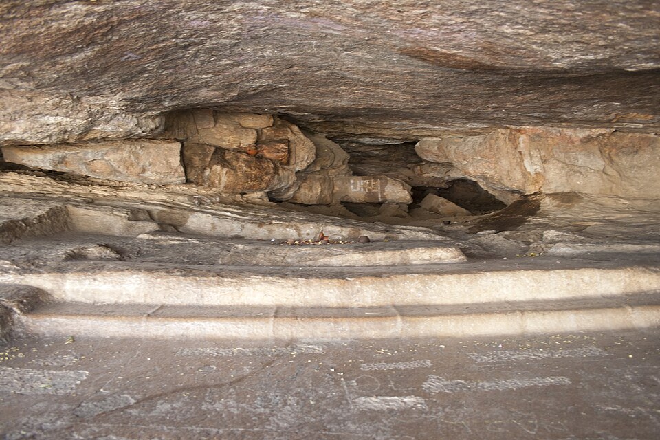 The Chera rock shelter at Pugalur: a natural overhang of grey-brown granite, a low dark cavity running back under the boulder, and in the foreground a smoothed rock brow and floor carrying rows of faint pale incised marks — the weathered Tamil-Brahmi lettering cut into the stone.