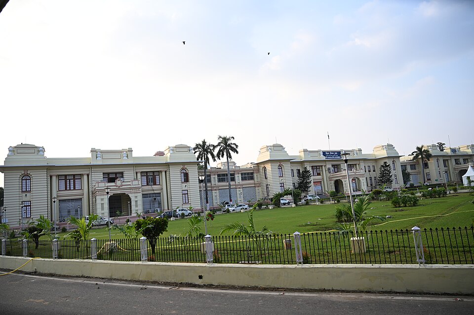 The long cream-coloured facade of the Bihar Vidhan Sabha in Patna, a low colonial-era block with arched windows, projecting bays and small domed roof pavilions, seen across a clipped green lawn with palms, a black iron fence and a whitewashed kerb wall along the road in front.