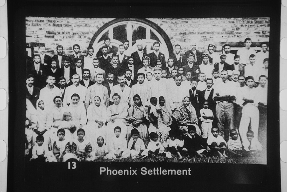 An old black-and-white lantern-slide frame, sprocket holes still visible down both edges, captioned 'Phoenix Settlement' along the bottom: some eighty men, women and children of the settlement posed in four rows in front of a stone building with a large round-headed window, the men in dark suits, waistcoats and caps behind, women in white saris seated in the middle row and small children cross-legged on the grass in front.