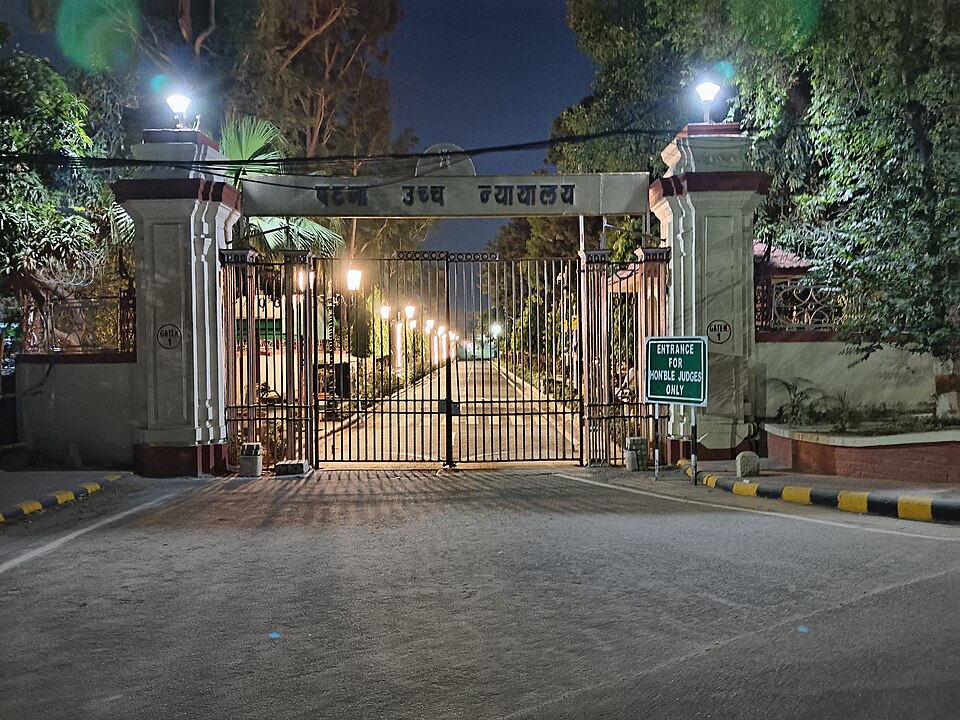 Gate No. 1 of the Patna High Court at night: a pale masonry gateway with 'पटना उच्च न्यायालय' lettered across its lintel, closed iron gates below, a green sign reading 'ENTRANCE FOR HON'BLE JUDGES ONLY' on the right pier, and a lamp-lit avenue receding into the dark court compound behind.