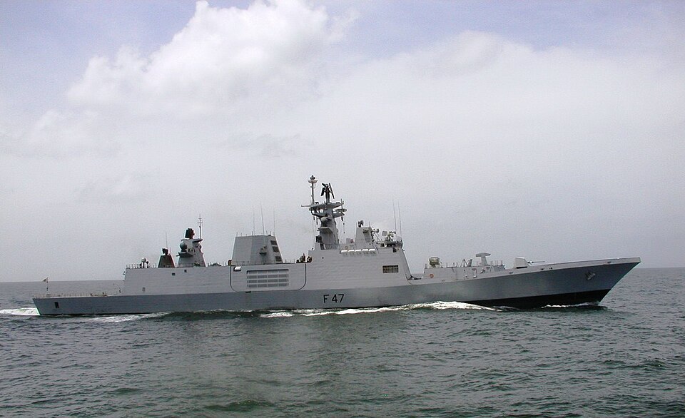 A grey Indian Navy frigate under way in a calm green sea, seen full broadside from starboard: sloped stealth-shaped hull and superstructure, pennant number F 47 painted amidships, a gun turret forward, a lattice mast carrying radar arrays and a domed sensor, and a low white bow wave curling away under a bank of cloud.
