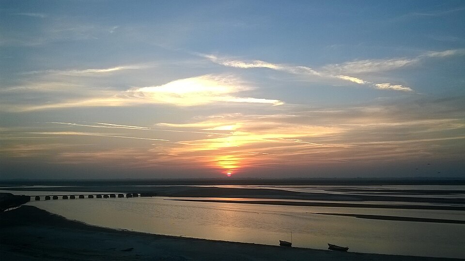 Sunset over the Ghaghara near Bahraich: the sun a red disc just above a flat, featureless horizon, its light laid down a wide sheet of still water that is broken up by long pale sandbars into several separate channels, a low many-arched causeway crossing the left of the frame, and two small wooden boats drawn up on the empty sand in the foreground.
