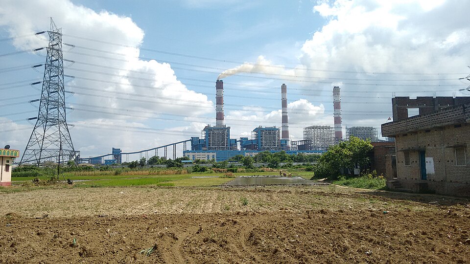 The Barh thermal power station seen across farmland: three tall chimneys banded red and white rise behind a row of blue steel boiler houses and a long covered coal conveyor, the leftmost stack trailing a white plume downwind, with a steel transmission tower and its cables at the left, a green paddy field and a ploughed brown plot in front, and an unplastered brick house at the right edge.