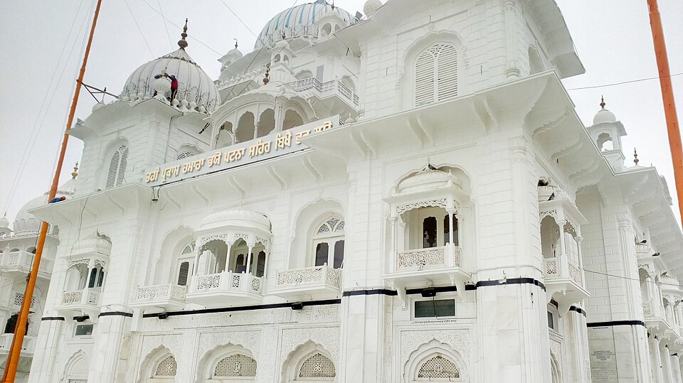 The white marble front of Takht Sri Patna Sahib photographed from below on a hazy day: tiers of arched openings, jharokha balconies and pierced latticework, a corner turret and two ribbed domes above, and a long gilt-lettered Gurmukhi inscription band running across the facade at first-floor level, with the orange nishan sahib mast at the left edge.