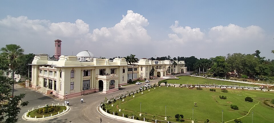 पटना में बिहार विधानसभा परिसर का ऊपर से लिया गया पूरा दृश्य: गुलाबी किनारों वाले मेहराबदार बरामदों और त्रिकोणी छज्जों वाली लंबी दोमंज़िली क्रीम रंग की इमारत, उसके बायें सिरे पर लाल ईंट का घड़ी-मीनार और पीछे एक नीचा चाँदी-सा गुंबद, दोनों ओर पीछे तक जाते खंड, बीच में घुमावदार तारकोल का रास्ता जिस पर कुछ कारें खड़ी हैं, और सामने कटे हुए झाड़ों तथा लैंप-पोस्टों वाला बड़ा छँटा हुआ लॉन; ऊपर ढेर-से सफ़ेद बादलों वाला नीला आकाश।